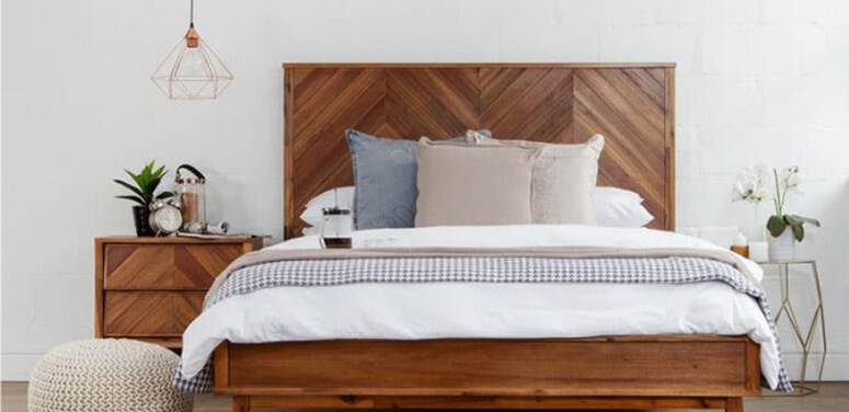 Neat, neutral coloured bedroom showing wooden bed and white bedding with grey and brown accent tones in throw and scatter cushions as well as  one wooden pedestal and another thin, gold metal pedestal